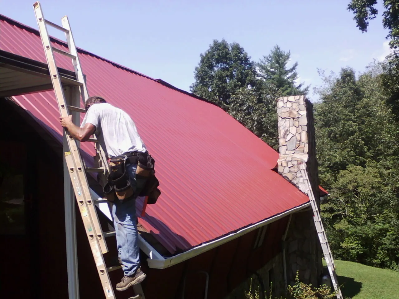 Roofer on a ladder beside a red metal roof and stone chimney — Soffit & Fascia Repair by Roofing Brothers in Tujunga